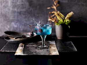 Photograph of a blue cocktail with a red cherry splashing into the cocktail